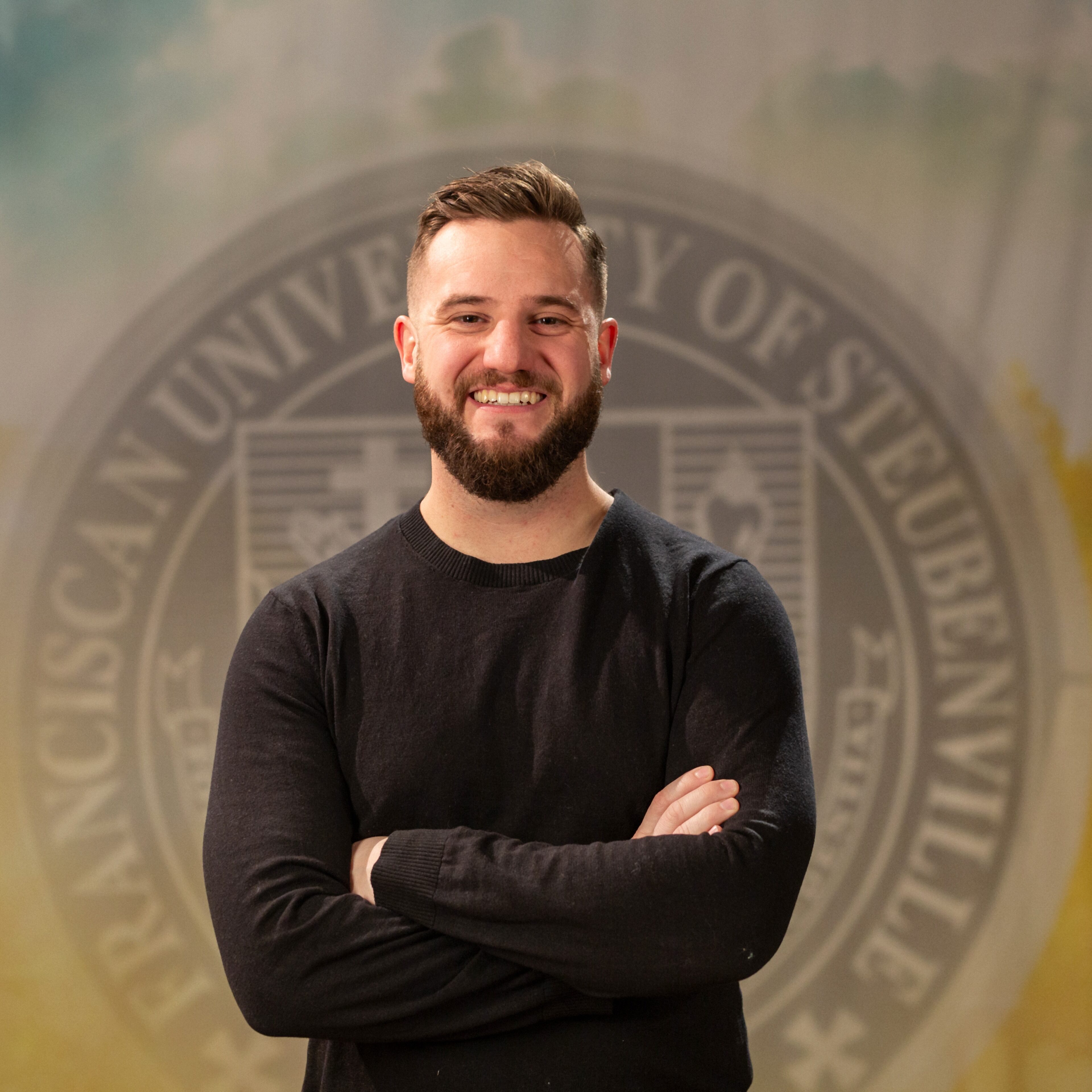 Portrait of John Paul Von Arx standing in front of the Franciscan University of Steubenville seal.